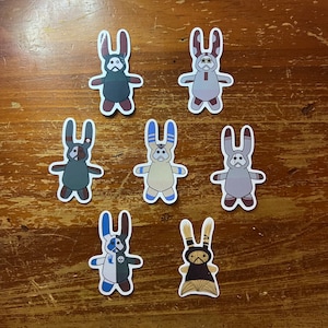 May include: Seven die-cut stickers featuring stylized rabbit-like figures with long ears. Each figure has a unique color scheme and pattern, including shades of gray, blue, tan, and black. The designs include various facial markings and symbols.