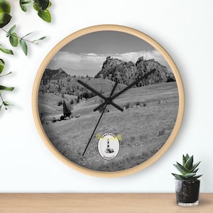 May include: Black and white clock with a wooden frame and a landscape image of a mountain range. The clock face has black hands and a small logo with the text "TIMOTHEOX" and a lighthouse graphic.