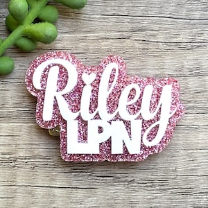 May include: A custom badge reel featuring a pink glitter background with the white text Riley and LPN layered on top. The name Riley is written in a cursive script font with a small heart above the letter i, while LPN is in block letters.
