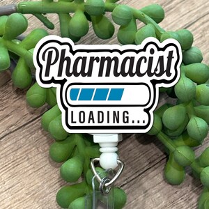 May include: A white badge reel featuring a black and white graphic with the text Pharmacist and a blue loading bar graphic above the text LOADING... in black. The badge reel has a clear plastic strap with a metal snap and a metal split ring.