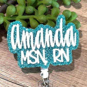 May include: A teal glitter badge reel featuring the white text Amanda MSN, RN in a script and block font combination. The badge reel has a white plastic base with a metal clip and a retractable cord mechanism for holding identification cards.
