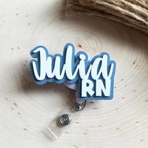 May include: A custom badge reel featuring a layered design with a dusty blue base and white lettering that reads Julia RN. The name Julia is written in a cursive script font, while the letters RN are in a bold, block sans-serif typeface.