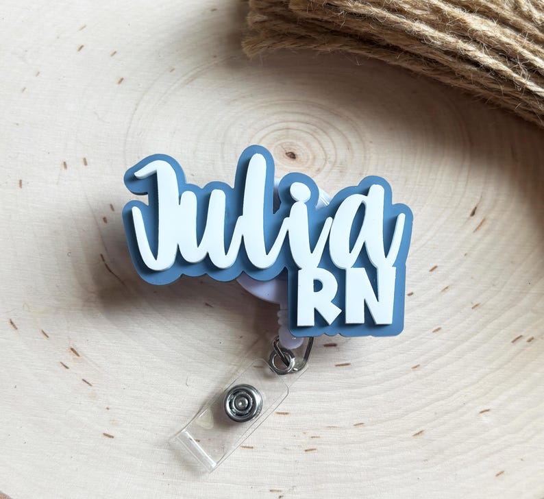May include: A custom badge reel featuring a layered design with a dusty blue base and white lettering that reads Julia RN. The name Julia is written in a cursive script font, while the letters RN are in a bold, block sans-serif typeface.