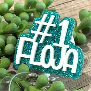 May include: A teal glitter badge reel featuring the white text #1 FLOJA in a bold, raised font. The badge reel has a circular plastic clip mechanism at the bottom, designed for holding identification cards or keys securely in place.