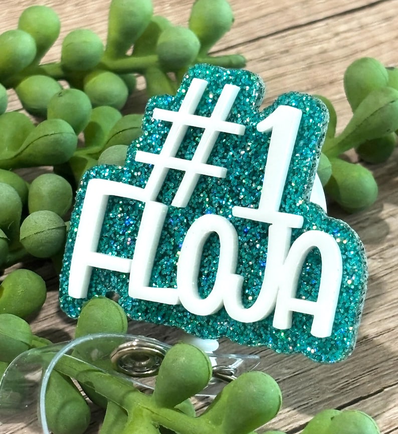 May include: A teal glitter badge reel featuring the white text #1 FLOJA in a bold, raised font. The badge reel has a circular plastic clip mechanism at the bottom, designed for holding identification cards or keys securely in place.