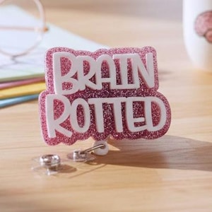 May include: A badge reel featuring a pink glittery background with the white text BRAIN ROTTED in a bold, layered font. The badge reel has a retractable cord mechanism with a clear plastic strap and a metal snap button at the bottom for attachment.