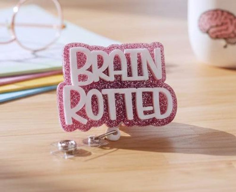 May include: A badge reel featuring a pink glittery background with the white text BRAIN ROTTED in a bold, layered font. The badge reel has a retractable cord mechanism with a clear plastic strap and a metal snap button at the bottom for attachment.