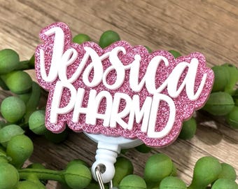May include: A custom badge reel featuring a pink glitter background with the white raised text Jessica PHARMD. The design is layered with a script font for the name and a bold sans-serif font for the professional title, attached to a white base.