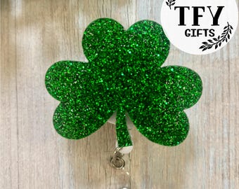 May include: Green glitter shamrock shaped badge holder with a clear retractable cord and a silver clip. The shamrock is attached to a white backing. The text "TFY GIFTS" is on a white circle with a black wreath design.