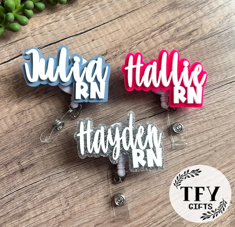 May include: Three custom badge reels featuring names Julia, Hallie, and Hayden in white script lettering with the letters RN in block font. The name plates are in light blue, hot pink, and silver glitter, each attached to a retractable cord.
