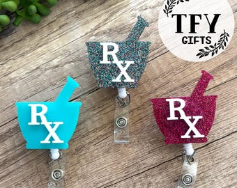 May include: Three badge reels featuring mortar and pestle shapes with the text Rx. One is solid turquoise, one is teal glitter, and one is magenta glitter. Each has a white Rx symbol and a clear plastic strap with a metal snap button.
