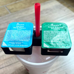 May include: Two small condiment cups with lids, one green and one blue, are attached to a black plastic holder. The cups are labeled "Garden Herb Ranch Sauce" and "Sweet & Spicy Sriracha Sauce". A red straw is inserted through the holder.