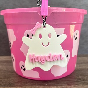 K&ouml;nnte beinhalten: Rosafarbener Plastik-Halloween-Eimer mit einem geisterf&ouml;rmigen Anh&auml;nger, auf dem "Hayden" in rosa Buchstaben steht. Der Eimer ist mit wei&szlig;en Geistern verziert.