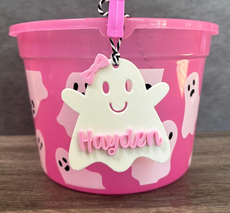 K&ouml;nnte beinhalten: Rosafarbener Plastik-Halloween-Eimer mit einem geisterf&ouml;rmigen Anh&auml;nger, auf dem "Hayden" in rosa Buchstaben steht. Der Eimer ist mit wei&szlig;en Geistern verziert.