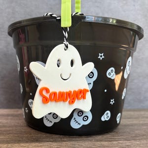 K&ouml;nnte beinhalten: Ein schwarzer Plastik-Halloween-Eimer mit einem wei&szlig;en, geisterf&ouml;rmigen Anh&auml;nger, auf dem "Sawyer" in orangefarbenen Buchstaben steht. Der Eimer ist mit wei&szlig;en Sch&auml;deln und Sternen verziert.