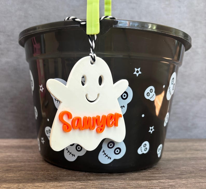 K&ouml;nnte beinhalten: Ein schwarzer Plastik-Halloween-Eimer mit einem wei&szlig;en, geisterf&ouml;rmigen Anh&auml;nger, auf dem "Sawyer" in orangefarbenen Buchstaben steht. Der Eimer ist mit wei&szlig;en Sch&auml;deln und Sternen verziert.