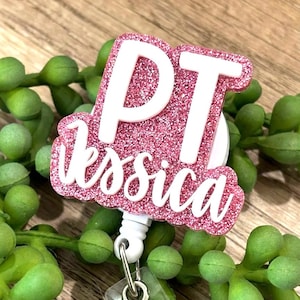 May include: A pink glitter badge reel with the text "PT Jessica" in white. The badge reel has a white retractable cord and a clear plastic clip. The background is a blurred image of green plant stems.
