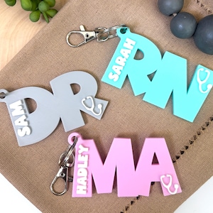 Könnte beinhalten: Drei personalisierte Schlüsselanhänger mit den Buchstaben "DR", "RN" und "MA" in verschiedenen Farben. Jeder Schlüsselanhänger hat einen Stethoskop-Anhänger und einen Namen darauf. Die Namen sind "Sam", "Sarah" und "Hadley".
