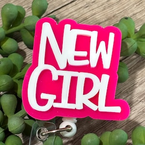 May include: A badge reel featuring a bright pink, die-cut acrylic topper with the white text NEW GIRL in a bold, sans-serif font. The topper is attached to a white plastic base with a retractable cord and a clear vinyl strap for identification.