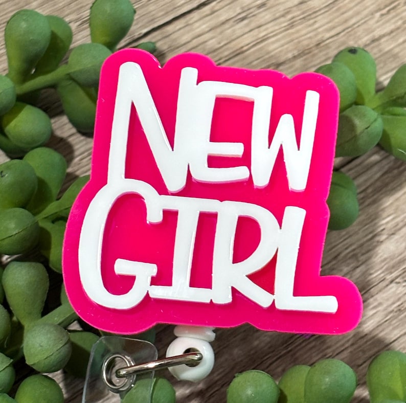 May include: A badge reel featuring a bright pink, die-cut acrylic topper with the white text NEW GIRL in a bold, sans-serif font. The topper is attached to a white plastic base with a retractable cord and a clear vinyl strap for identification.