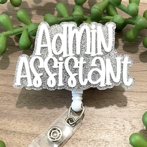 May include: A white and silver glitter badge reel with the words "Admin Assistant" in a bold, clear font. The badge reel has a retractable cord and a metal clip for attaching to clothing.