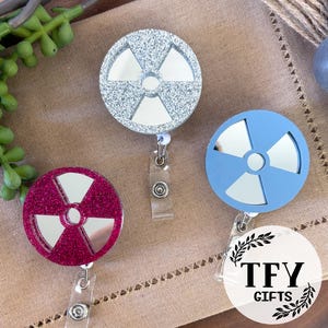 May include: Three circular badge reels featuring a radiation symbol design with mirrored cutouts. One reel is silver glitter, one is solid light blue, and one is magenta glitter. Each includes a clear plastic strap and a metal snap button closure.