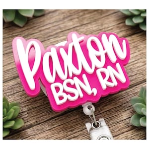 May include: A custom badge reel featuring a bright pink acrylic base with the name Paxton and the professional credentials BSN, RN in raised white script and block lettering. The badge reel includes a clear plastic strap and a metal clip.