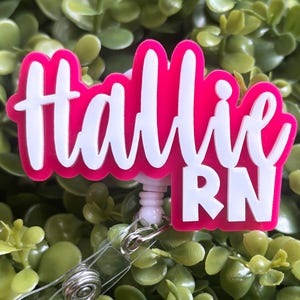 May include: A custom badge reel featuring a bright hot pink acrylic backing with the name Hallie and the letters RN layered in white script and block lettering. The design is attached to a retractable plastic cord mechanism with a metal clip.