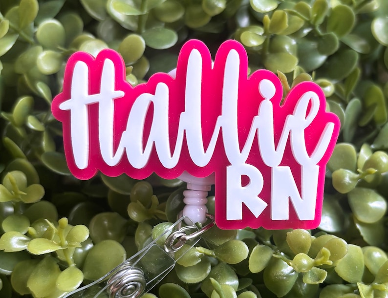 May include: A custom badge reel featuring a bright hot pink acrylic backing with the name Hallie and the letters RN layered in white script and block lettering. The design is attached to a retractable plastic cord mechanism with a metal clip.