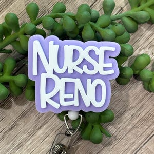 May include: A badge reel featuring a light purple, cloud-shaped acrylic topper with the white text NURSE RENO embossed on the front. The badge reel includes a clear plastic strap and a metal clip mechanism attached to the bottom of the topper.