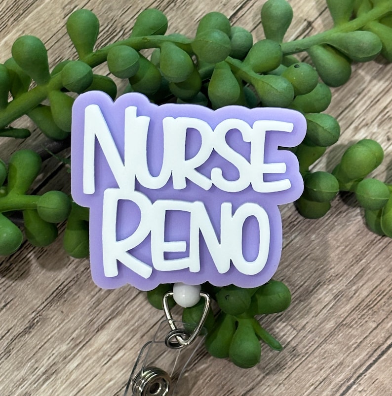 May include: A badge reel featuring a light purple, cloud-shaped acrylic topper with the white text NURSE RENO embossed on the front. The badge reel includes a clear plastic strap and a metal clip mechanism attached to the bottom of the topper.