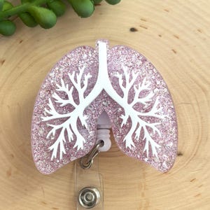 May include: A badge reel featuring a glittery light pink lung-shaped topper with a white anatomical bronchial tree design overlaid on the surface. The badge reel mechanism is white plastic with a metal clip attached to the bottom of the lung shape.