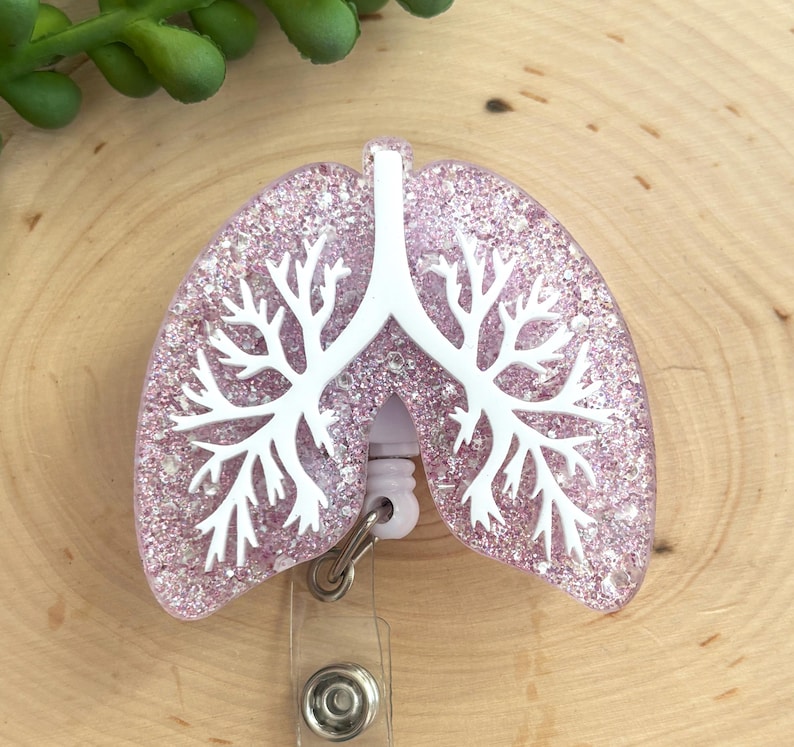 May include: A badge reel featuring a glittery light pink lung-shaped topper with a white anatomical bronchial tree design overlaid on the surface. The badge reel mechanism is white plastic with a metal clip attached to the bottom of the lung shape.