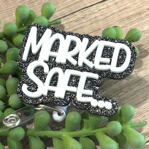 May include: A badge reel featuring a black glittery background with the white raised text "MARKED SAFE..." in a bold sans-serif font. The badge includes a clear plastic strap with a metal snap button attachment for securing identification cards.