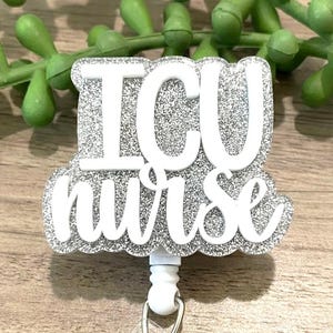 May include: A glittery silver and white badge reel with the words "ICU nurse" in white lettering. The badge reel has a white retractable cord and a metal clip. The background features green plant stems.