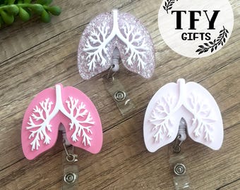May include: Three badge reels shaped like human lungs, each featuring a white bronchial tree design. One reel is light pink, one is solid white, and one is a translucent light purple with embedded glitter. Each has a clear plastic strap and metal snap.