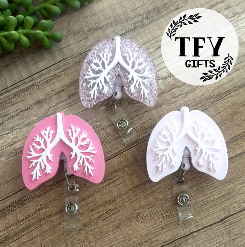 May include: Three badge reels shaped like human lungs, each featuring a white bronchial tree design. One reel is light pink, one is solid white, and one is a translucent light purple with embedded glitter. Each has a clear plastic strap and metal snap.