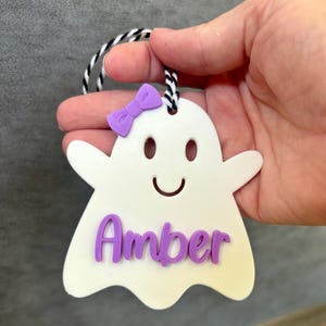 K&ouml;nnte beinhalten: Wei&szlig;er, geisterf&ouml;rmiger Anh&auml;nger mit einer lila Schleife und dem Namen "Amber" in lila Buchstaben.