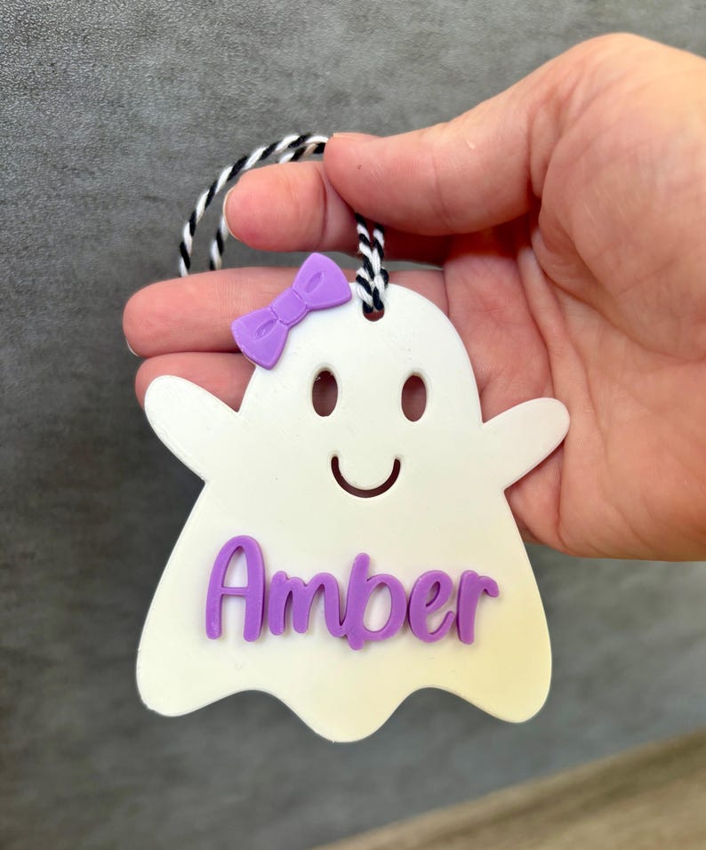 K&ouml;nnte beinhalten: Wei&szlig;er, geisterf&ouml;rmiger Anh&auml;nger mit einer lila Schleife und dem Namen "Amber" in lila Buchstaben.
