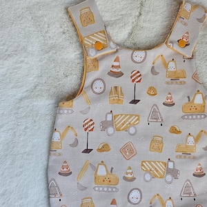 May include: Baby overalls in a tan fabric, patterned with construction vehicles, cones, and signs in yellow and brown. The overalls have yellow edging and button fastenings on the straps. The text "Little Digger" is printed on the straps.