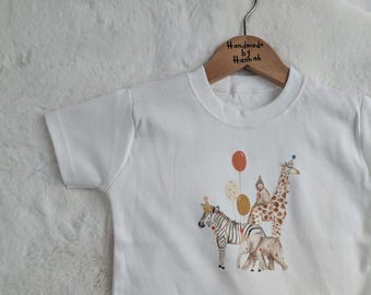 May include: White short-sleeved t-shirt featuring a colorful illustration of a zebra, elephant, giraffe, and monkey with balloons. The shirt is hanging on a wooden hanger that says "Handmade by Hannah".