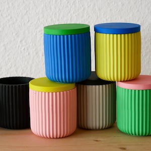 Könnte beinhalten: Ein Set aus sechs bunten zylindrischen Behältern mit geriffelten Seiten und passenden Deckeln. Die Behälter sind pyramidenförmig gestapelt und aus einem glatten, matten Material gefertigt. Die Farben sind blau, gelb, rosa, schwarz, weiß und grün.