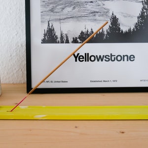 Könnte beinhalten: Ein gelber Räucherstäbchenhalter mit einem weißen marmorartigen Design. Ein Holz-Räucherstäbchen brennt und raucht. Der Räucherstäbchenhalter steht auf einer Holzoberfläche. Im Hintergrund befindet sich ein gerahmtes Schwarzweiß-Poster mit dem Text "Yellowstone" und Informationen über den Park.