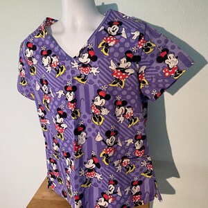 May include: A purple scrub top featuring an all-over pattern of Minnie Mouse in various poses. The short-sleeved top has a V-neck and two pockets. The Minnie Mouse characters are in red, black, white, and yellow. The background has a striped and polka dot design.