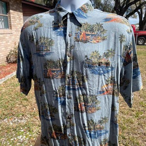 May include: Light blue button-down shirt with a tropical island print. The shirt features a pattern of palm trees, surfboards, and ocean scenes. It has short sleeves and a chest pocket. The shirt is displayed on a mannequin.