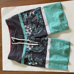 May include: Board shorts with a dark gray base, featuring a tropical floral pattern in teal, white, and gray. Teal and white stripes accent the sides. The shorts have a drawstring closure and a red interior lining.