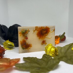 May include: A rectangular bar of clear resin with embedded orange and yellow dried flowers. Decorative fall-themed elements, including acorns and leaves, surround the bar. The bar is approximately 3 inches long.