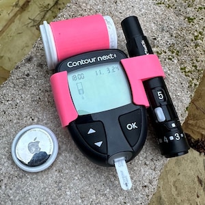 Könnte beinhalten: Ein rosa-schwarzes Blutzuckermessgerät mit einem digitalen Display, das die Zeit 18:00 und das Datum 11.3.24 anzeigt. Das Messgerät hat einen Knopf mit der Aufschrift "OK" und eine kleine Lanzette an der Seite. Ein schwarz-weißes Apple AirTag befindet sich im Vordergrund.