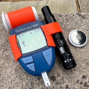 May include: A blue Contour Plus BLUE blood glucose meter with a reading of 16.8, a lancet, and a black lancet pen with a FastClix label. The meter is attached to a 3D printed orange holder.