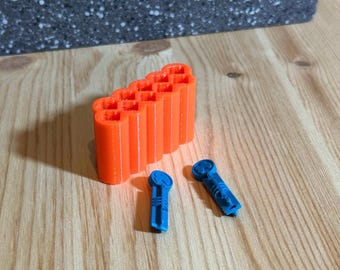 May include: An orange 3D-printed object with a grid-like top and several vertical columns, along with two blue objects. The items are on a light wood surface, with a dark gray textured block in the background. The objects appear to be components or parts.